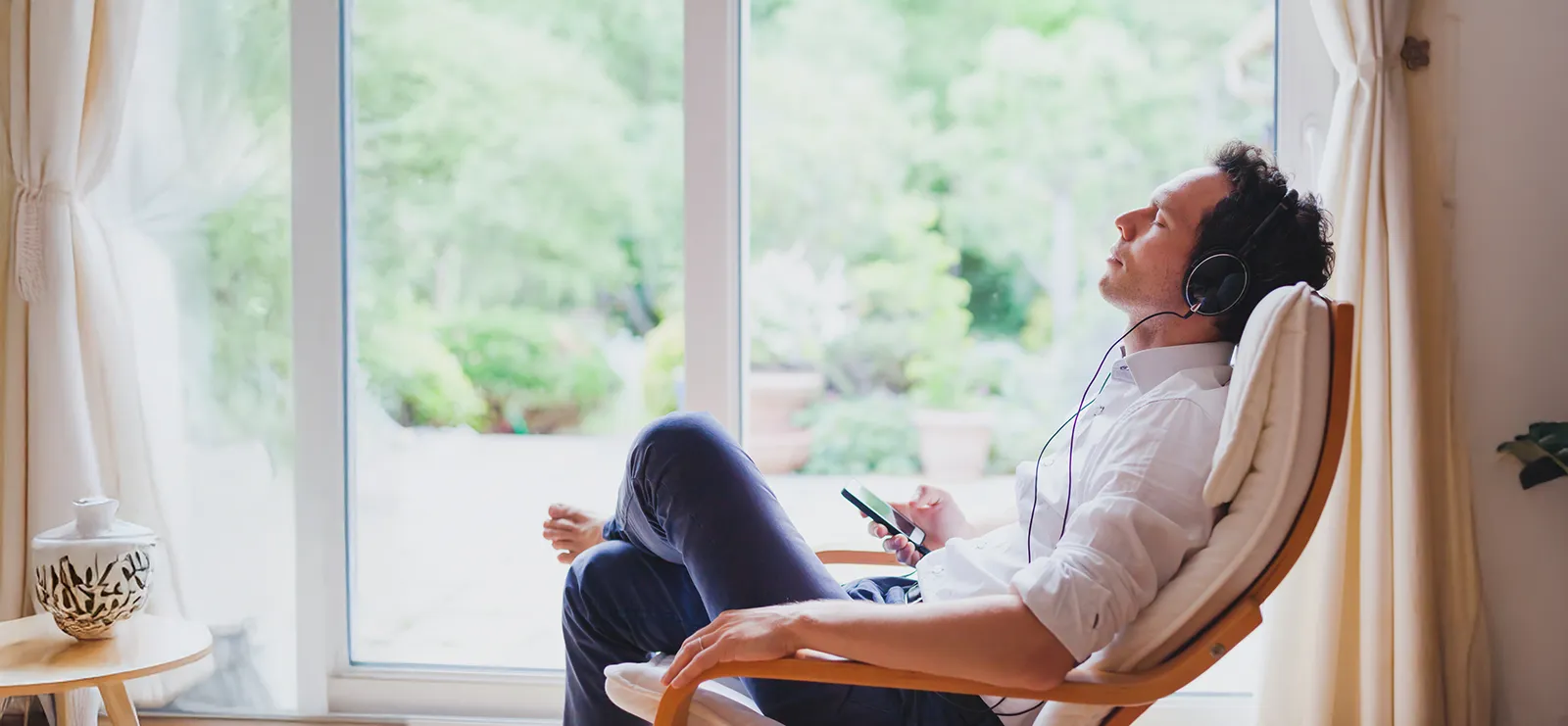 Homme détendu dans un fauteuil près d'une grande fenêtre, écoutant de la musique avec des écouteurs.