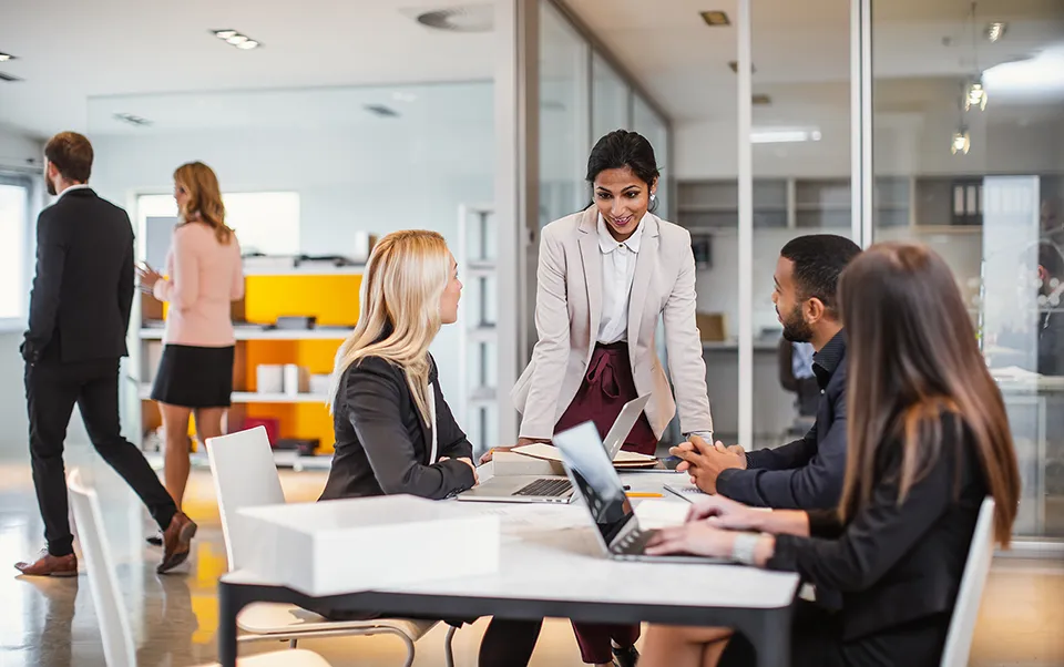 At a meeting in a well-lit conference room, a group of 4 people discuss graphs laid out on a table.