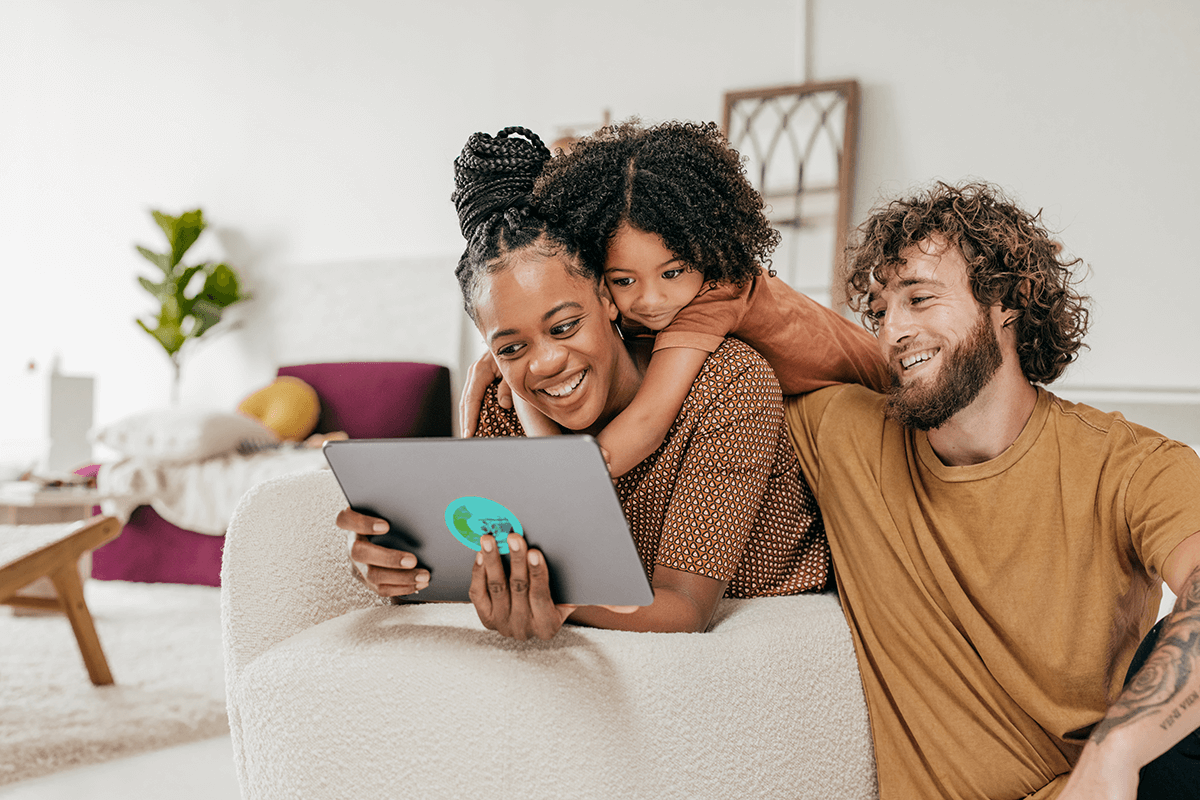 A mother, father and young daughter sit together in their living room. The mother leans on a chair and holds a tablet. The father and daughter huddle around her, all smiling while they watch the tablet.