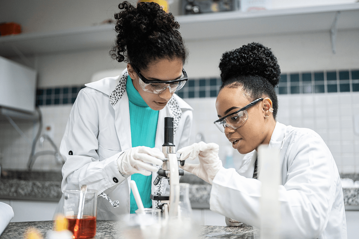 Two female students in lab coats and protective eyewear and gloves look into a microscope in a laboratory.