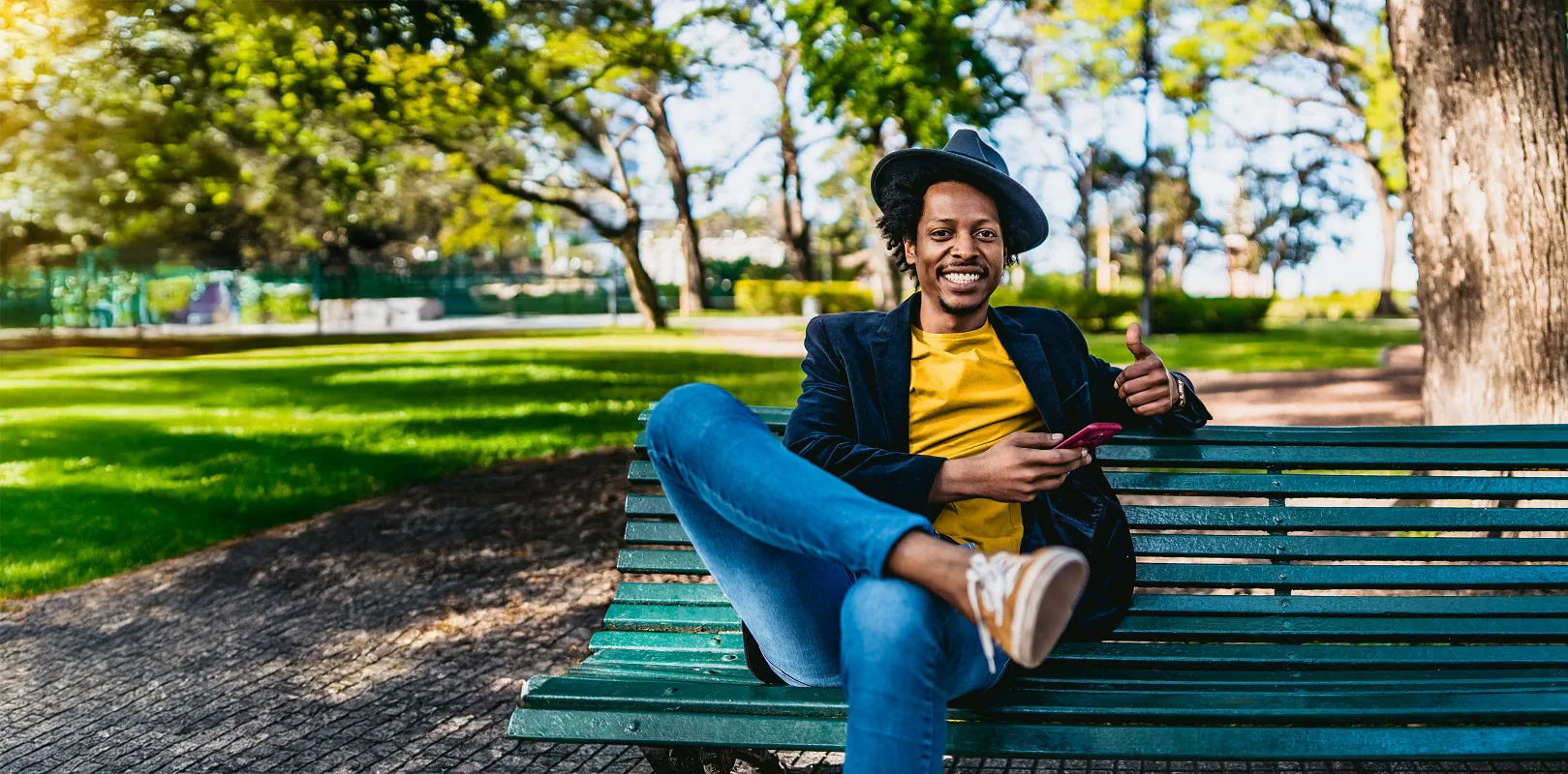 A stylish man sitting on a park bench holds a cell phone and gives a thumbs up.