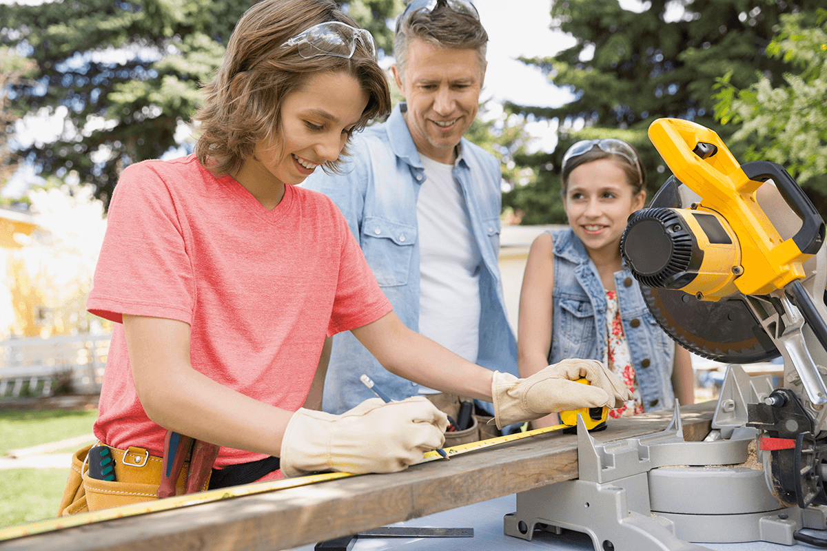 Une famille de trois travaille sur un projet de construction. La mère, qui porte des gants de travail, une ceinture à outils et des lunettes de protection sur la tête, mesure une pièce de bois sous le regard du père et de la fille.