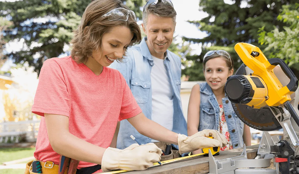 A family of three works on a construction project. The mother, wearing work gloves, a tool belt and protective eyewear perched on top of her head, measures a piece of wood while the father and daughter look on.