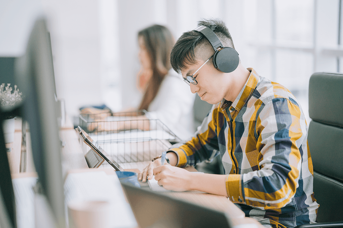 Un jeune homme est assis à un bureau partagé devant un écran d'ordinateur. Il porte des écouteurs et écrit dans un cahier. L'autre étudiant en arrière-plan semble également étudier.