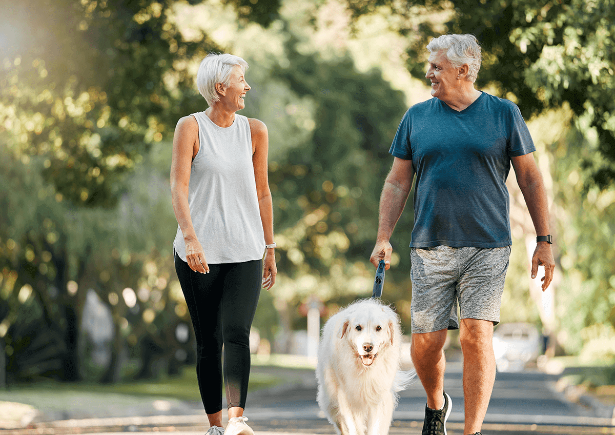 Two seniors smile at each other while leisurely walking with their dog.