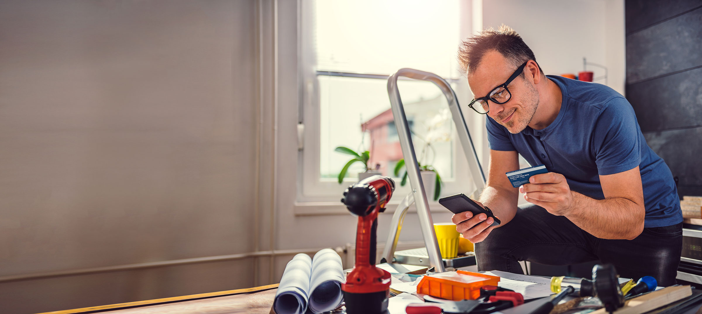 A man in glasses stands beside a ladder resting one foot on a rung. There’s a drill and other supplies on the table in front of him. His elbow rests on his knee while holding his mobile phone. In his other hand he holds a bank card. He smiles while looking at his phone.