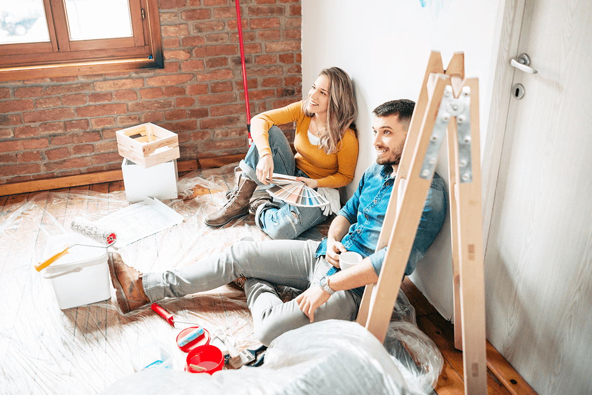 A smiling couple sit on the floor of a room with a plastic drop sheet down. Around them are a ladder and painting supplies. The woman holds fanned out paint swatches in her hand while he holds a coffee mug. 