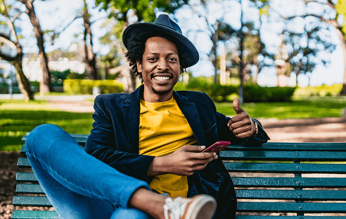 A young man wearing a fedora hat sits outside on a park bench, holding his phone. He smiles at the camera and gives a thumbs up.