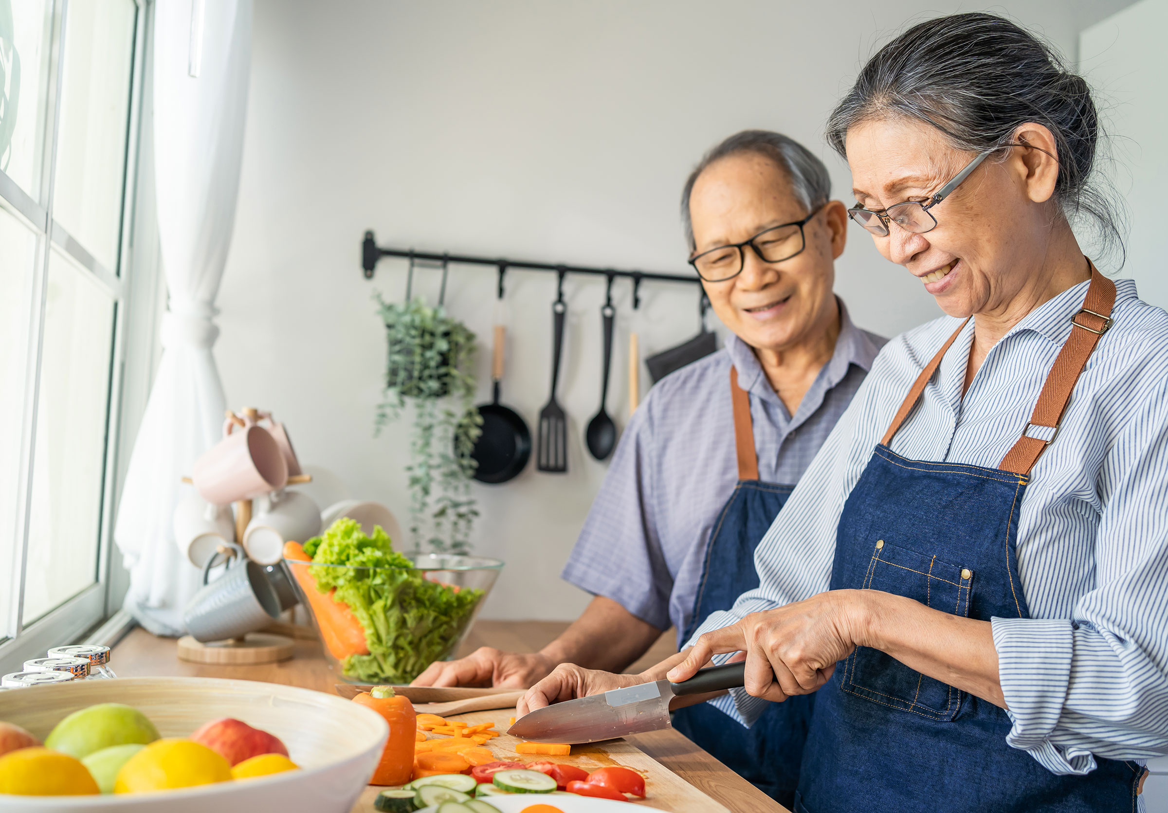 A smiling retired couple stand at a kitchen counter, each wearing an apron. The woman is holding a knife and cutting vegetables.