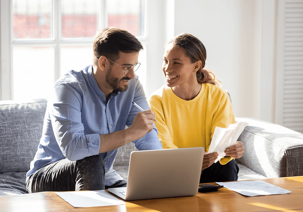 A couple sit together on a couch with a laptop, papers and a calculator on the table in front of them.