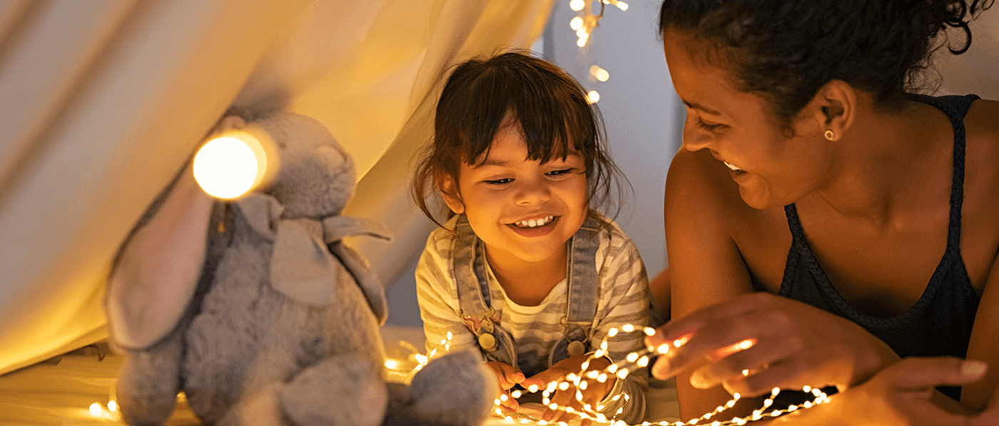 A mother and daughter are in a cloth tent set up indoors. Surrounded by soft fairy lights, they lay happily on their stomachs, propped up on their arms.