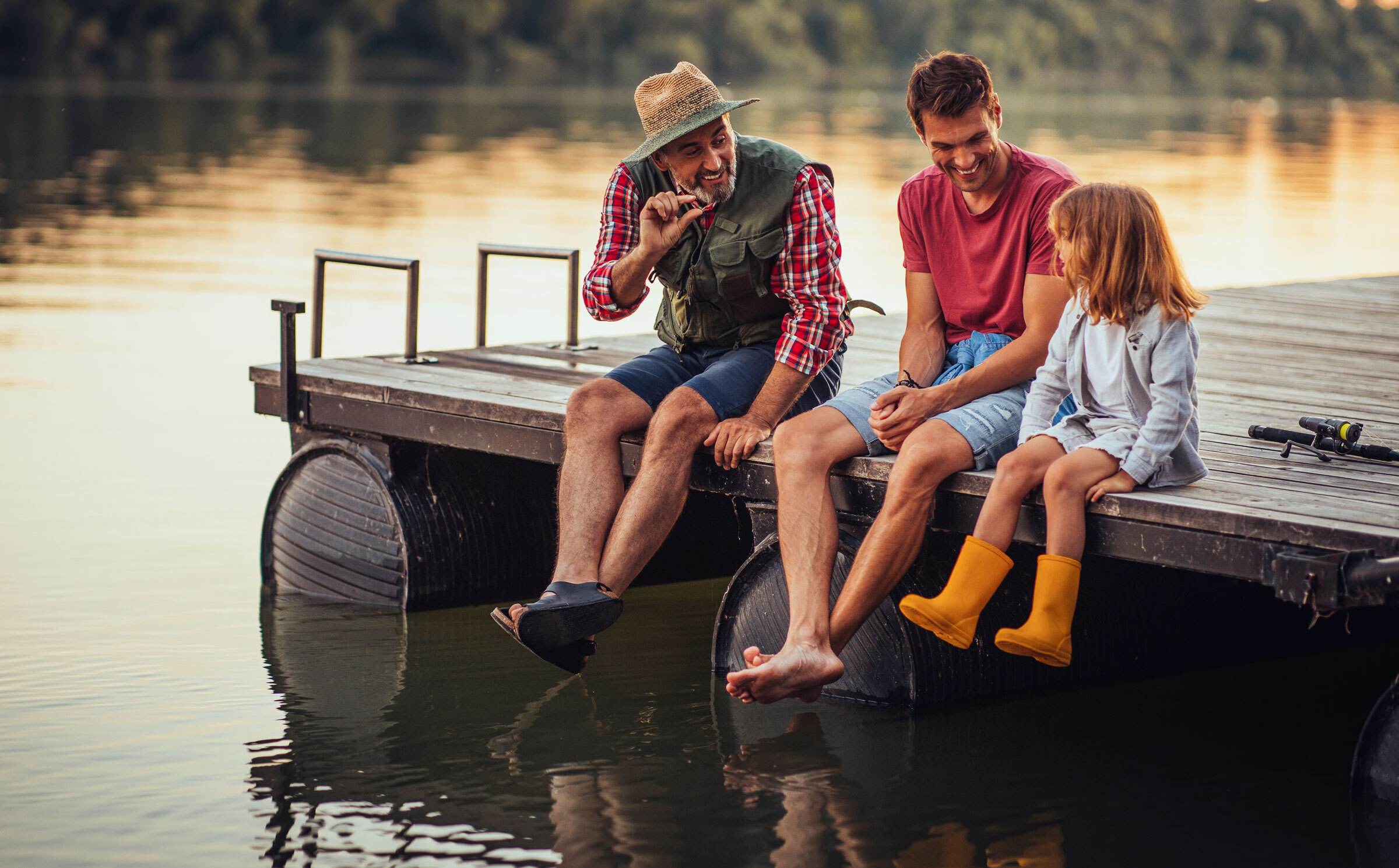 A grandfather, father and daughter sit on a dock with their legs dangling over the water.
