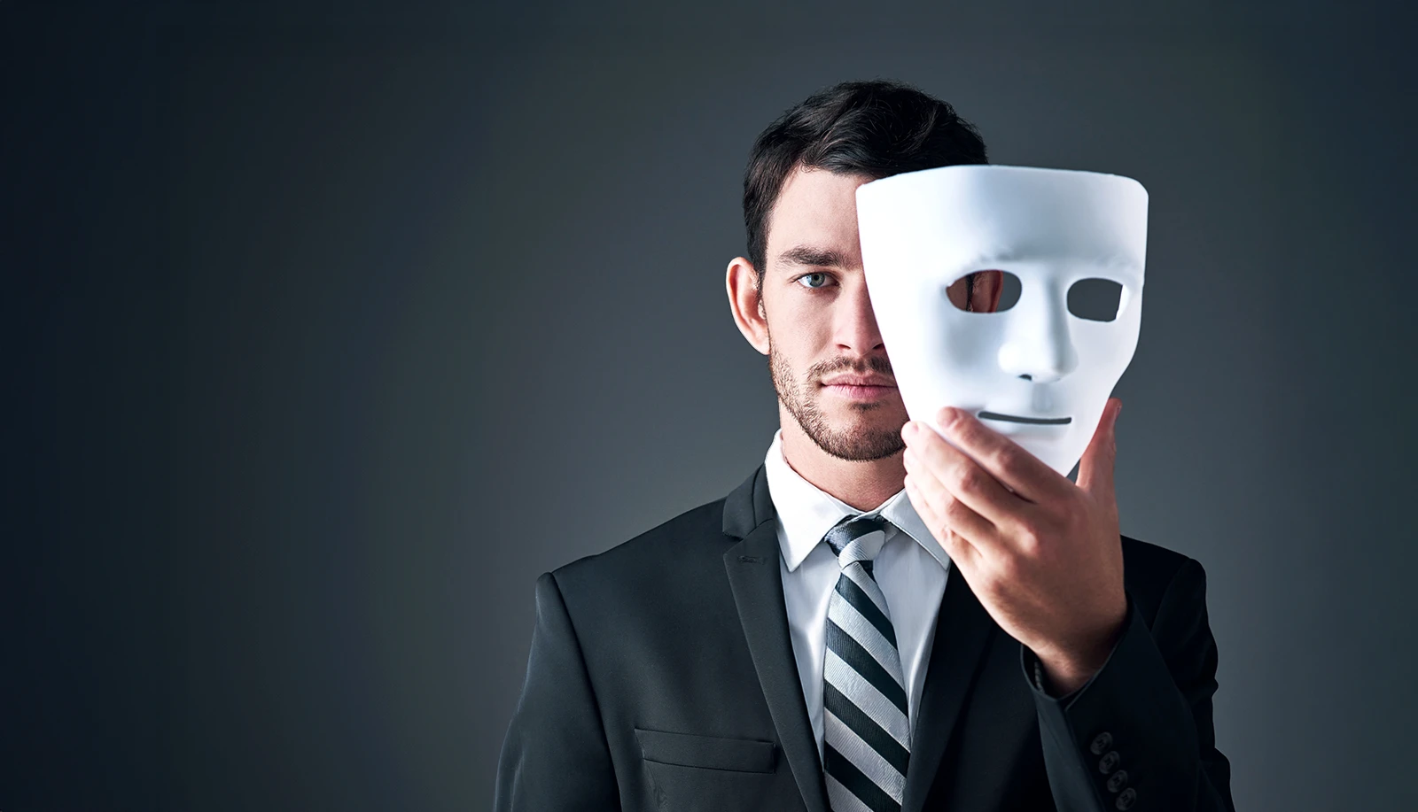 Un homme en complet-cravate tient un masque blanc qui couvre la moitié de son visage.