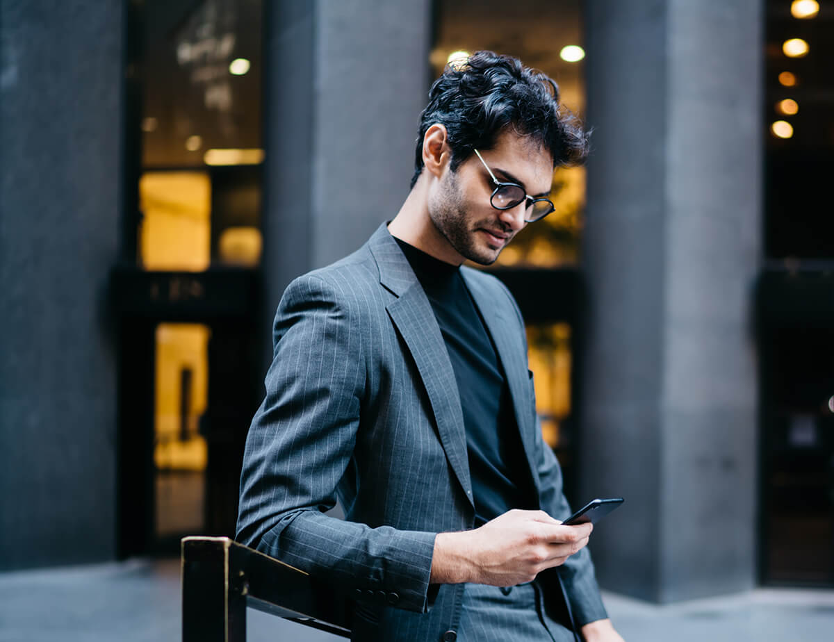 Un homme se tient à l'extérieur d'un grand immeuble à bureaux. Il regarde son téléphone tout en s'appuyant sur une rampe en métal.