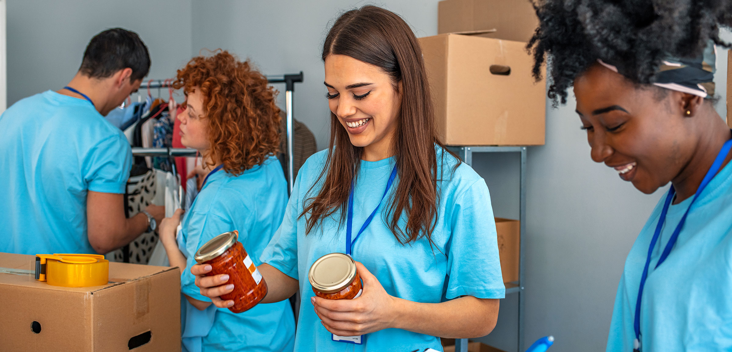 A group of smiling volunteers wearing matching blue shirts organize and sort canned food donations into boxes.