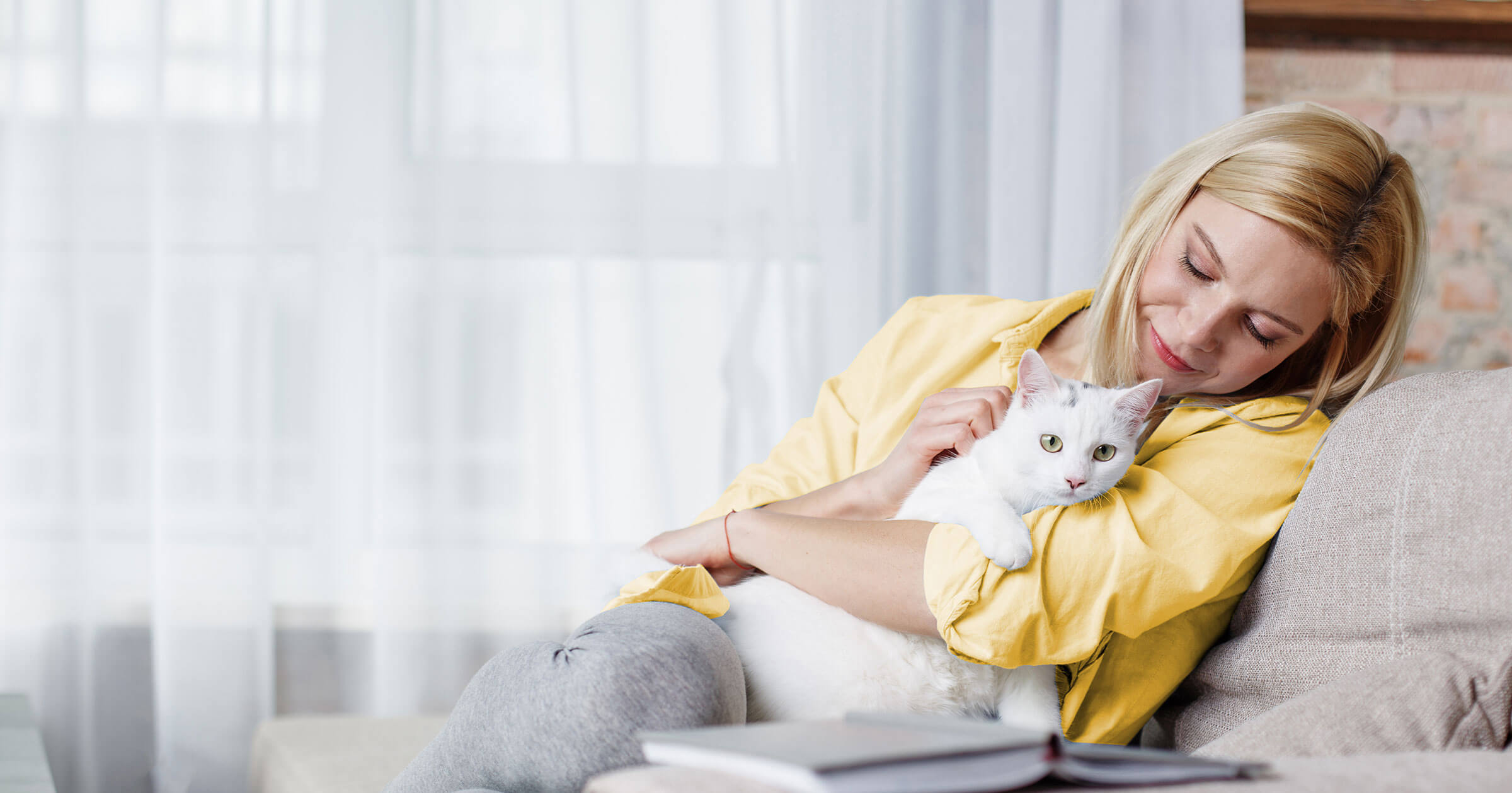 A woman wearing a yellow shirt sits on a couch holding a white cat.