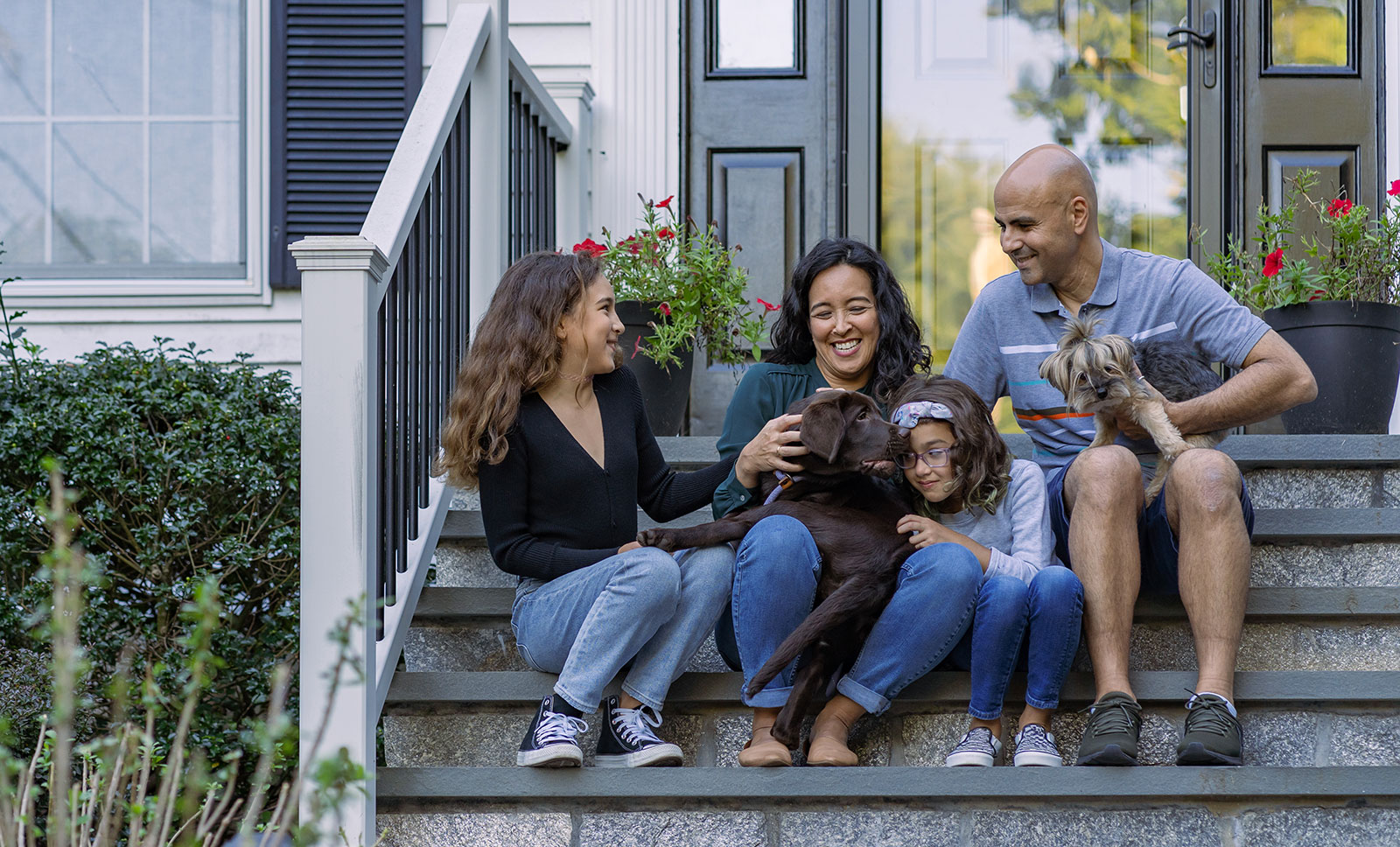 Une famille souriante est assise dans les escaliers devant une maison, accompagnée de deux chiens.