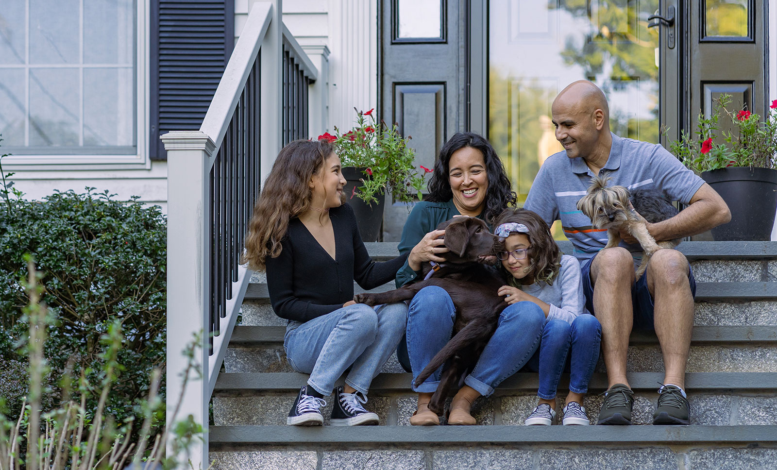 A family of four smile and laugh while playing with their two dogs on the stairs in front of their home.