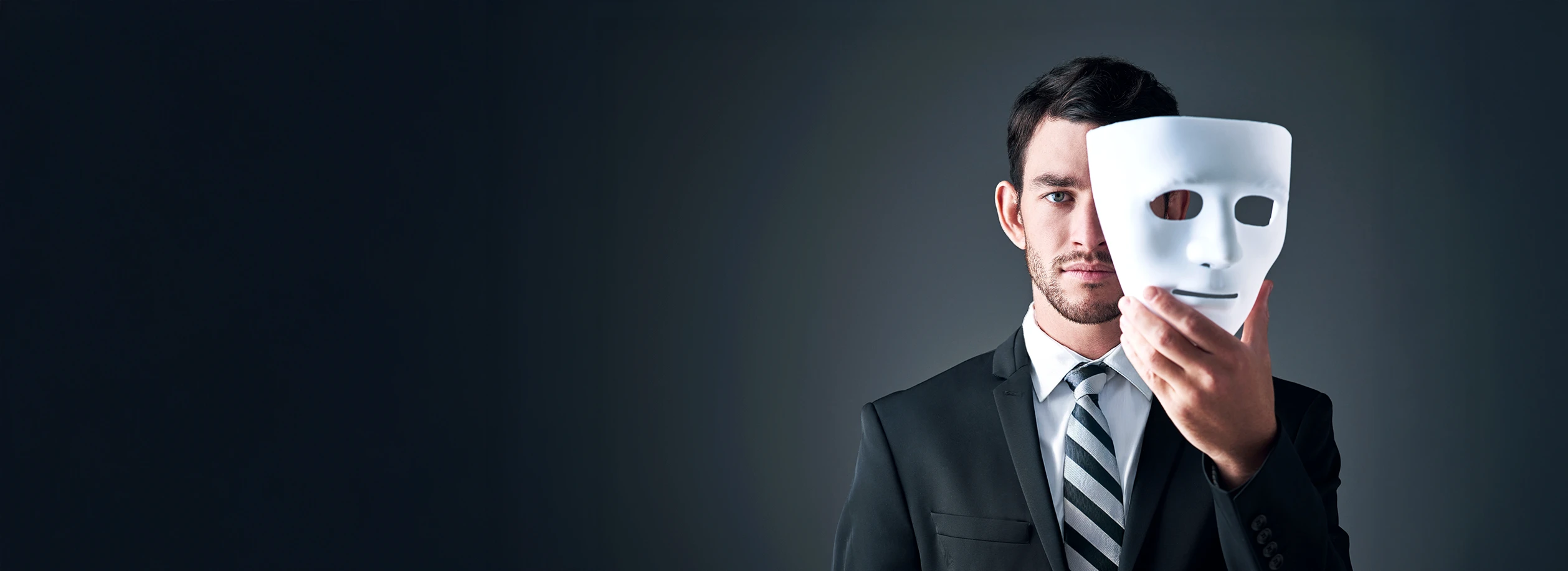 A man wearing a suit and tie holds a white mask covering half of his face.