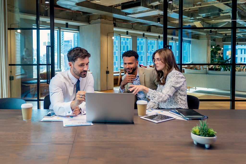 Three office employees talk in front of a laptop computer.