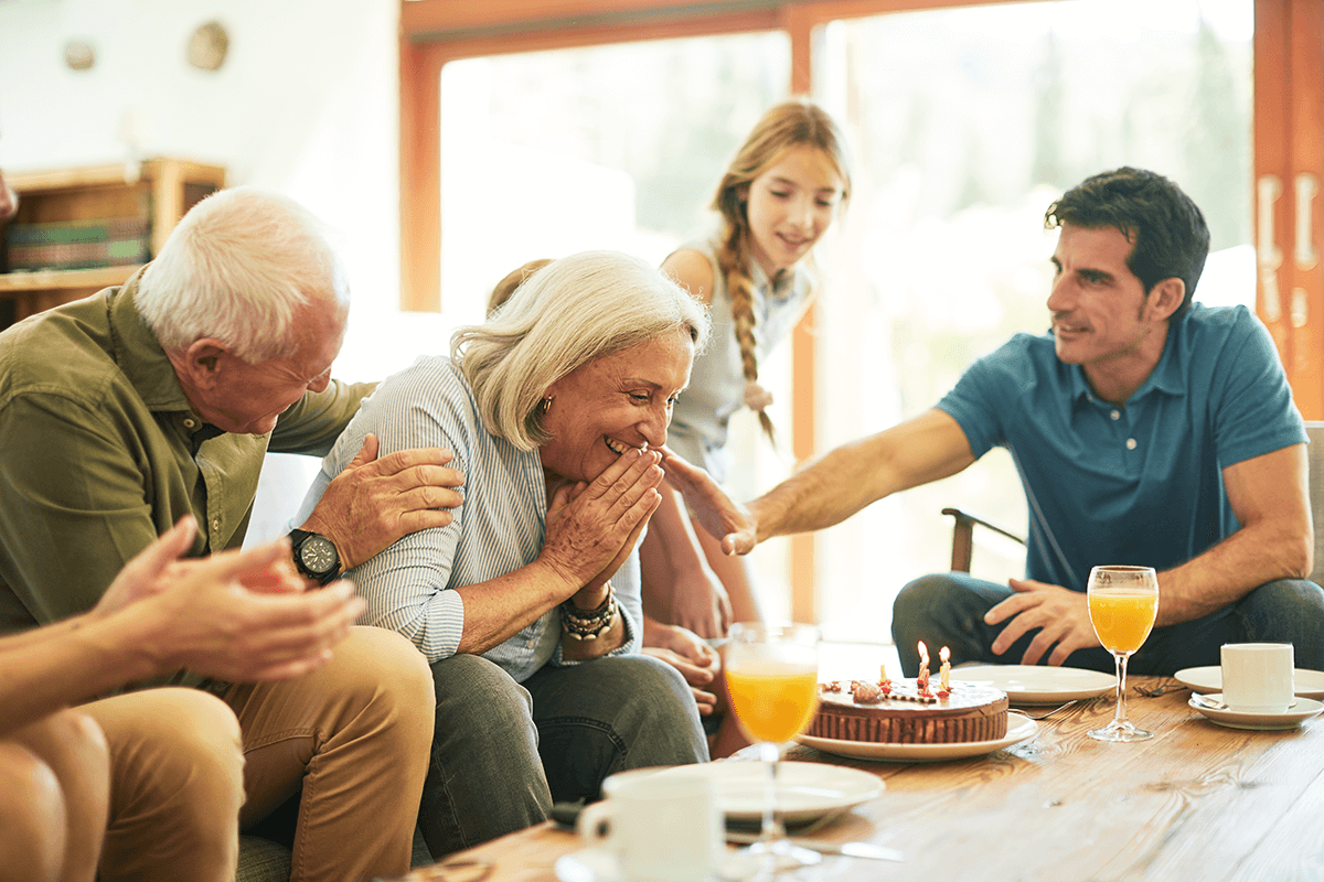 A senior woman smiles with joy as her family surrounds her with a surprise cake, celebrating a special occasion together.