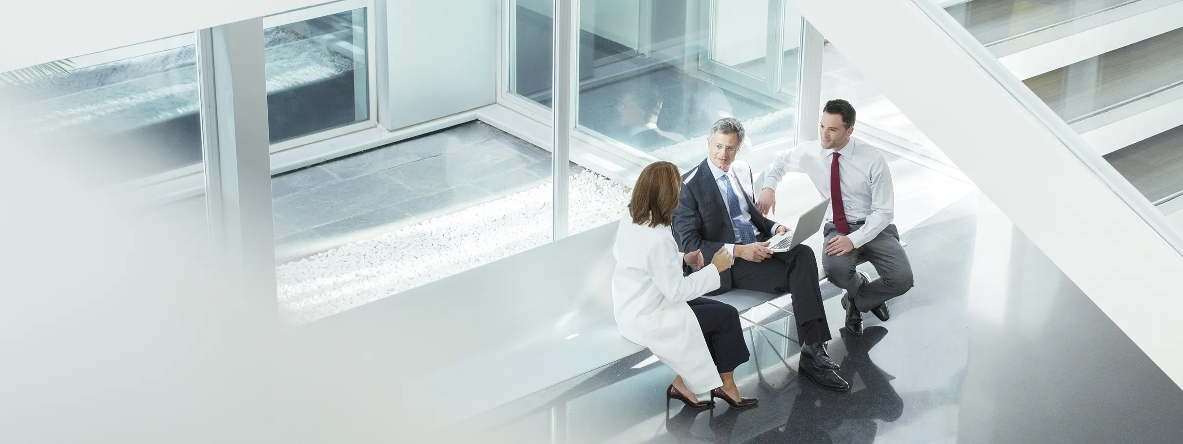 Three business professionals sit on a bench in a building lobby, chatting.