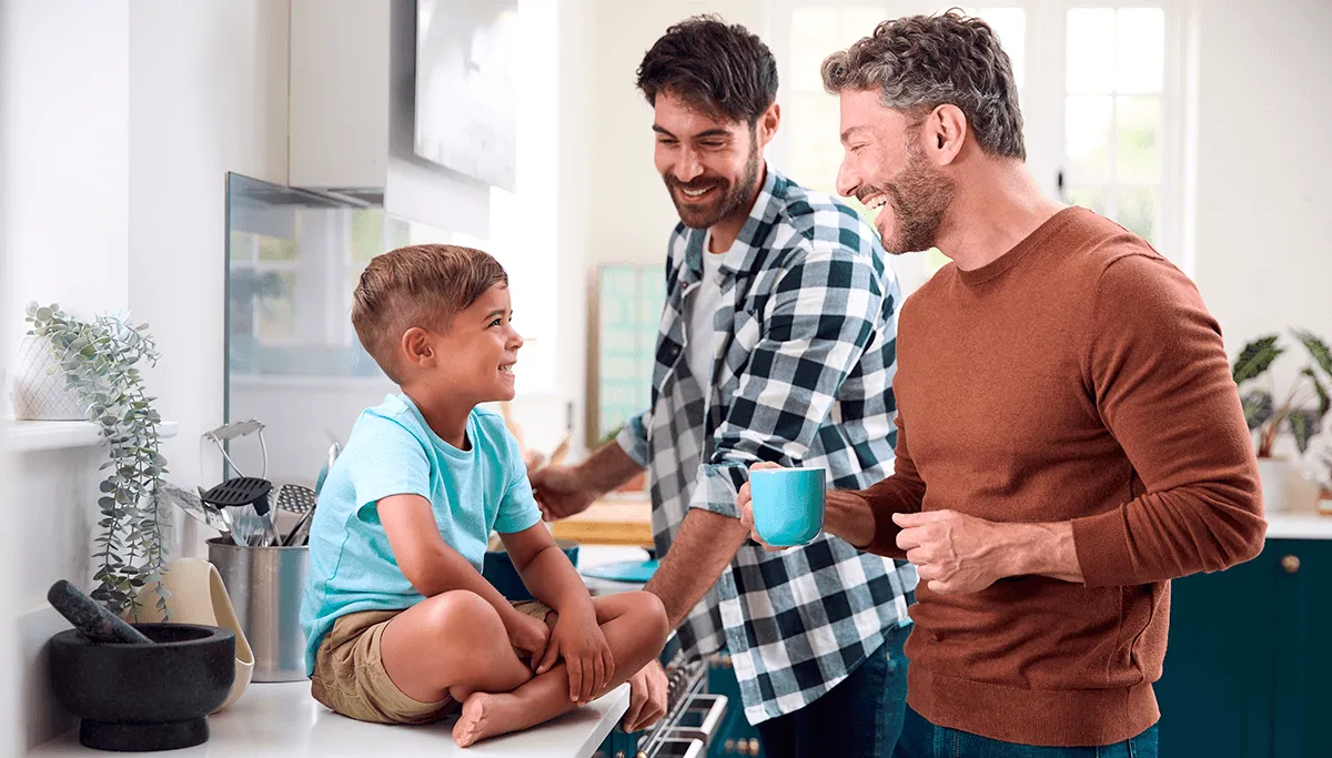 Dans une cuisine moderne et lumineuse, deux pères sourient à leur jeune garçon assis les jambes croisées sur le comptoir.