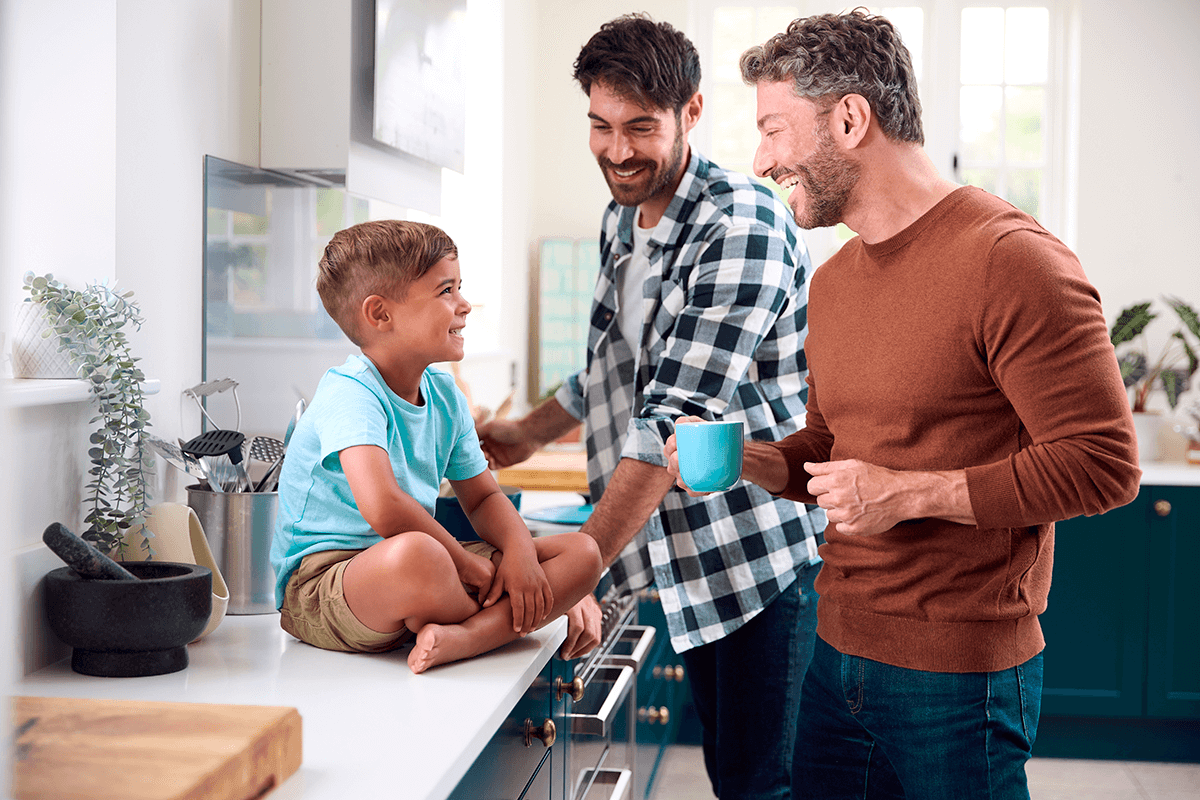 Dans une cuisine moderne et lumineuse, deux pères sourient à leur jeune garçon assis les jambes croisées sur le comptoir.
