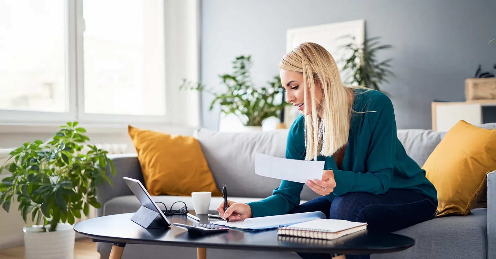 Sitting on a couch in a living room, a woman leans over a coffee table with pen and paper in hand. A notebook, calculator and tablet are spread out on the table.