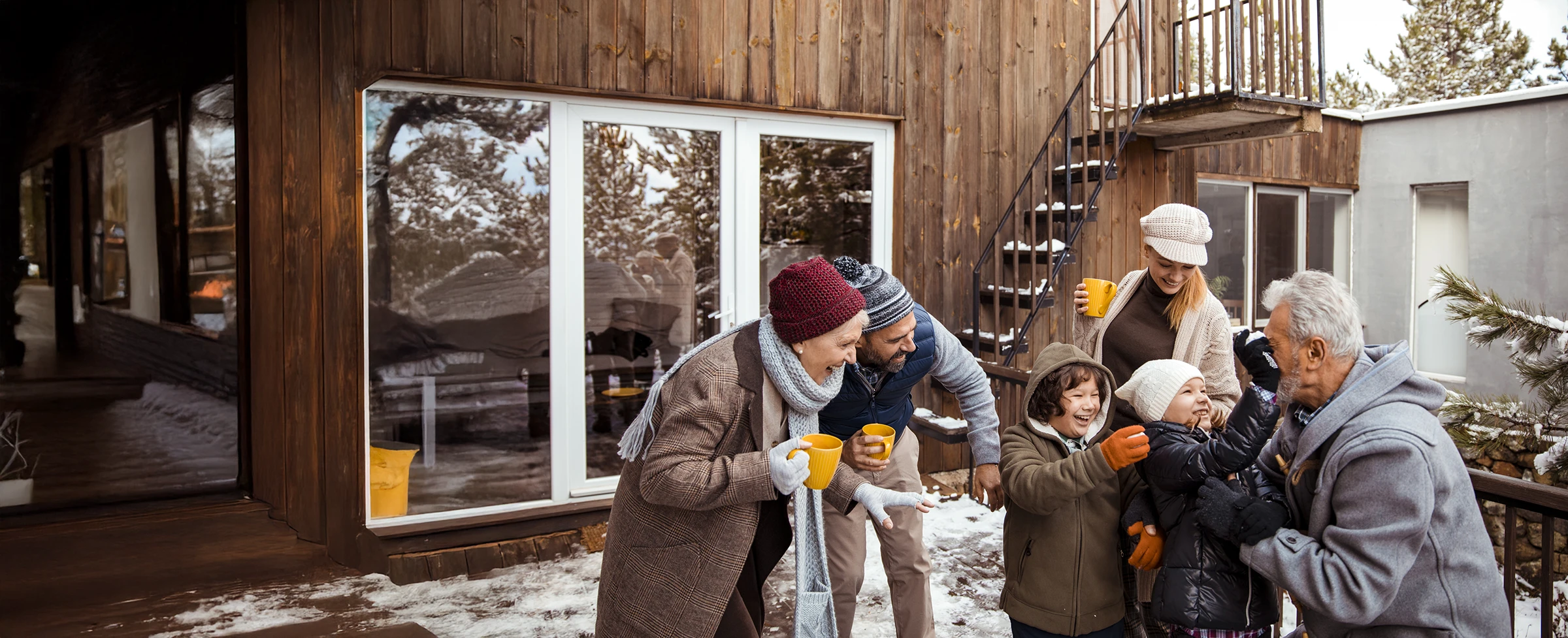 Outside a home on a winter’s day, an intergenerational family of 6 play with snowballs and sip hot drinks.
