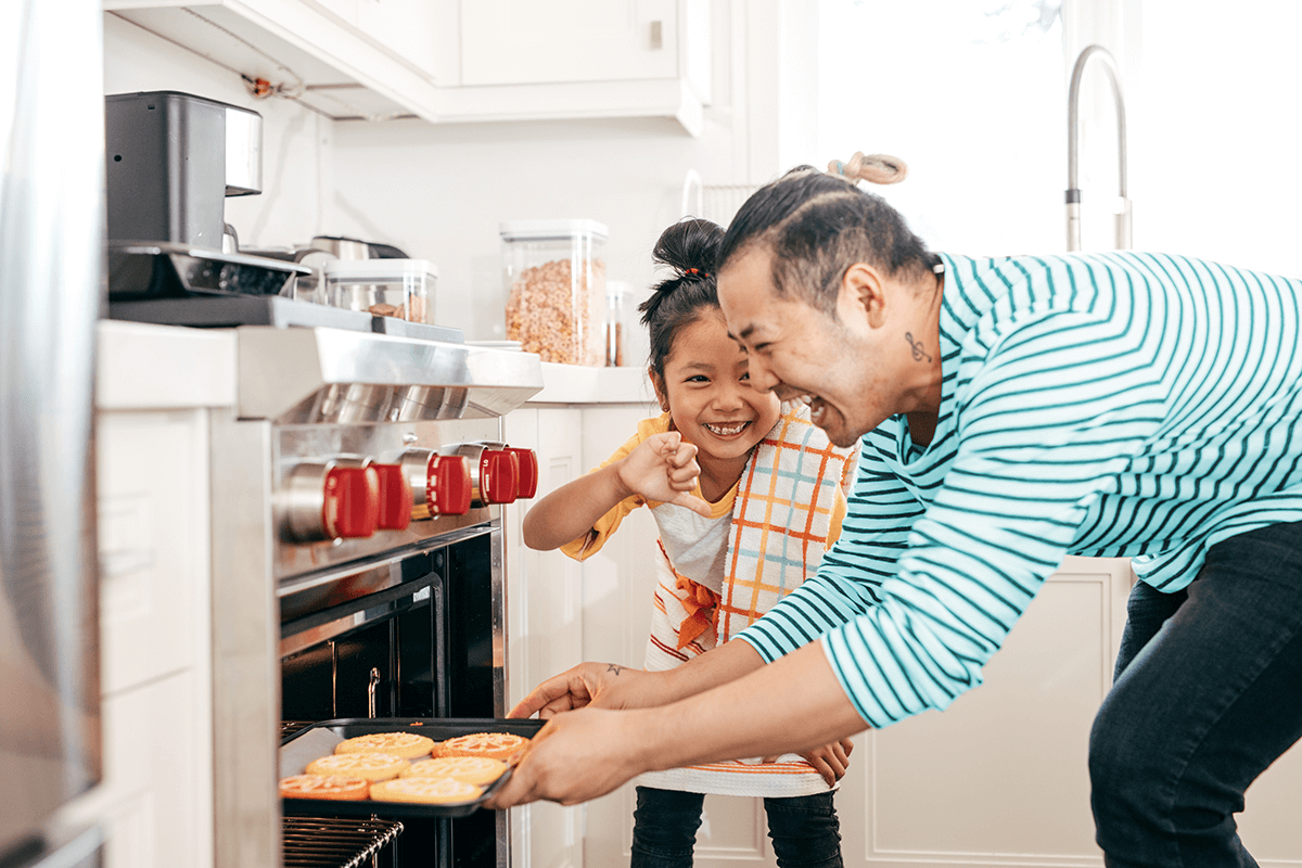 A father and daughter bend over in front of an oven. The father laughs while pulling freshly baked cookies out of the oven. The daughter smiles and gives him a thumbs down.