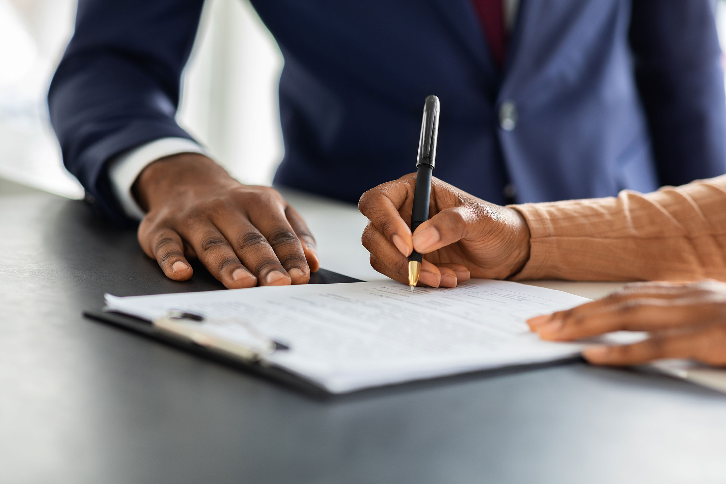 The hand of a person holds a pen and signs a document, while the hand of a man wearing a suit sits on the desk beside the papers.