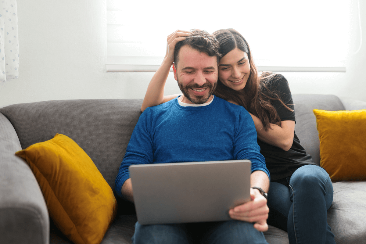 A happy couple sits closely together on a couch. They’re both looking at the laptop on the man’s lap.