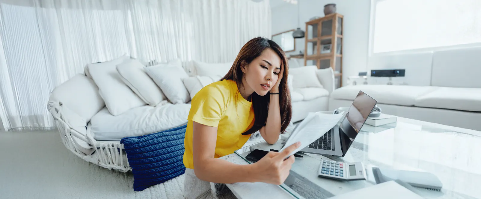 Une femme est assise par terre devant la table basse de son salon. Elle est penchée sur un ordinateur portable et une calculatrice, des papiers à la main.
