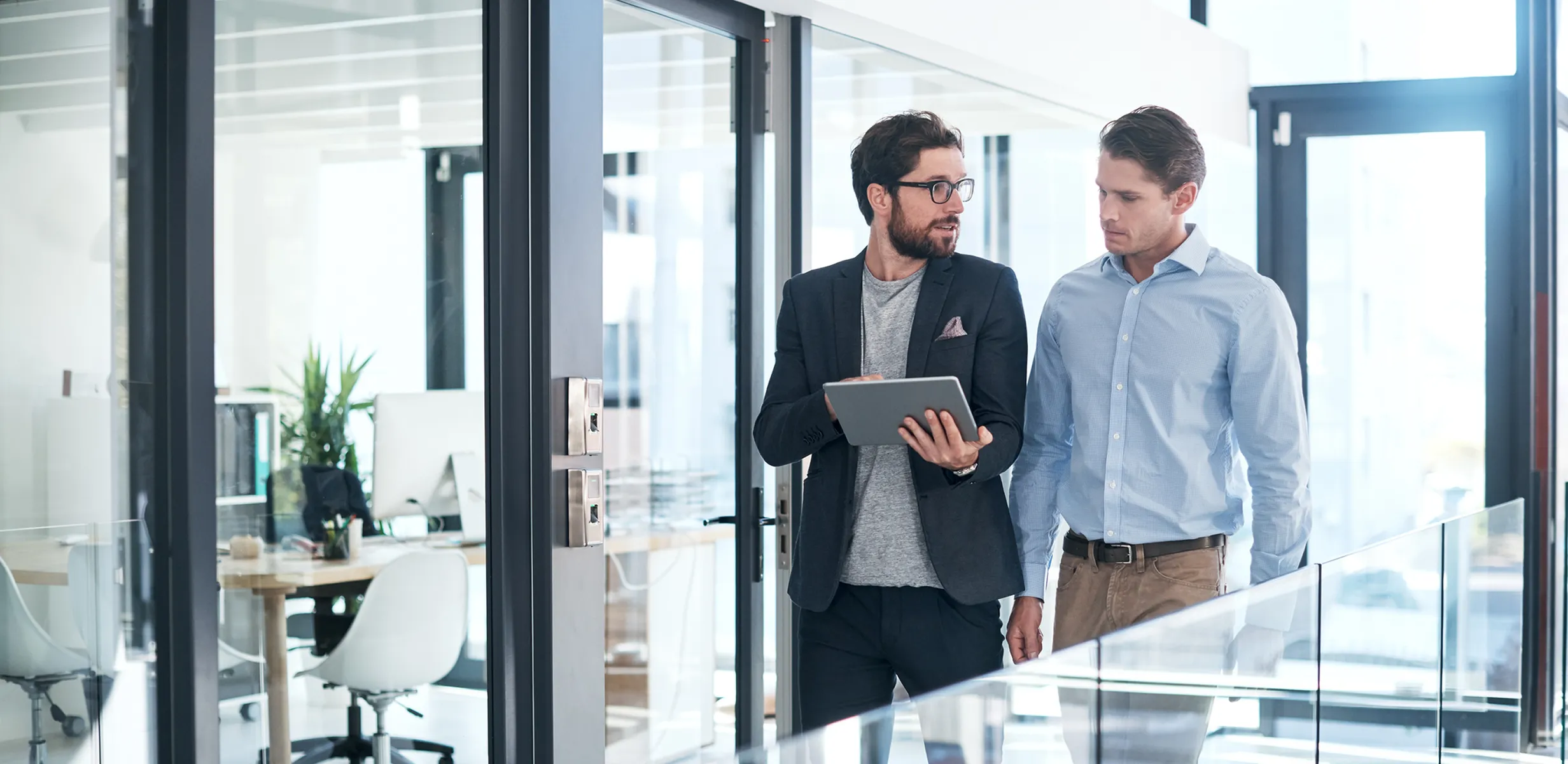 Two men in an office walk and talk while looking at a tablet.