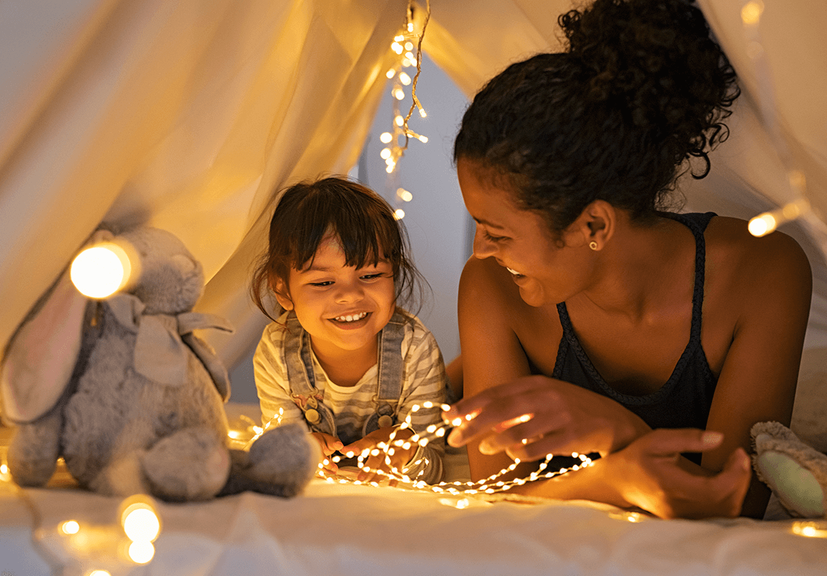 Une mère et sa fille se trouvent dans une tente en tissu installée à l'intérieur. Entourées de douces lumières féeriques, elles sont allongées sur le ventre, appuyées sur leurs bras.