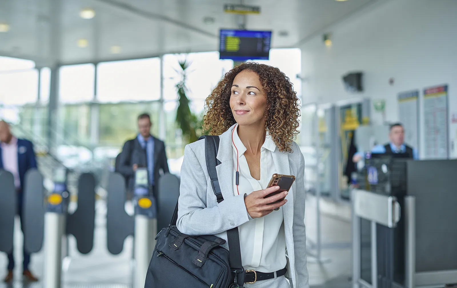 A woman wearing a business suit holds her cell phone in a transit station, a computer bag slung over her shoulder.