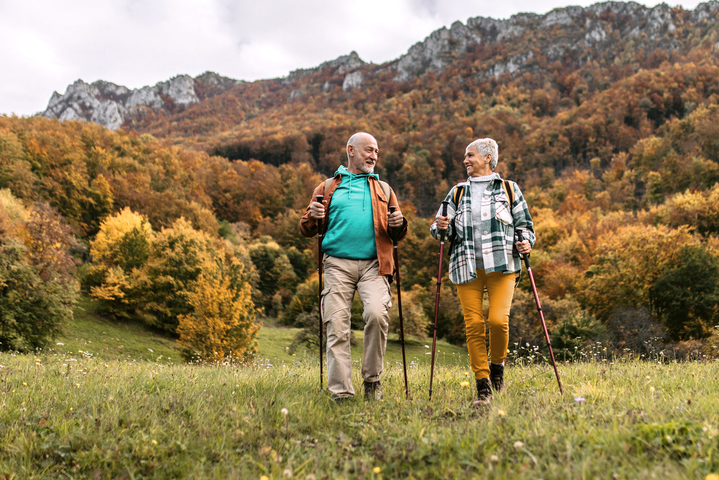 A retired couple hike with walking poles through a grassy field. Behind them is a mountain range and trees covered in fall leaves.