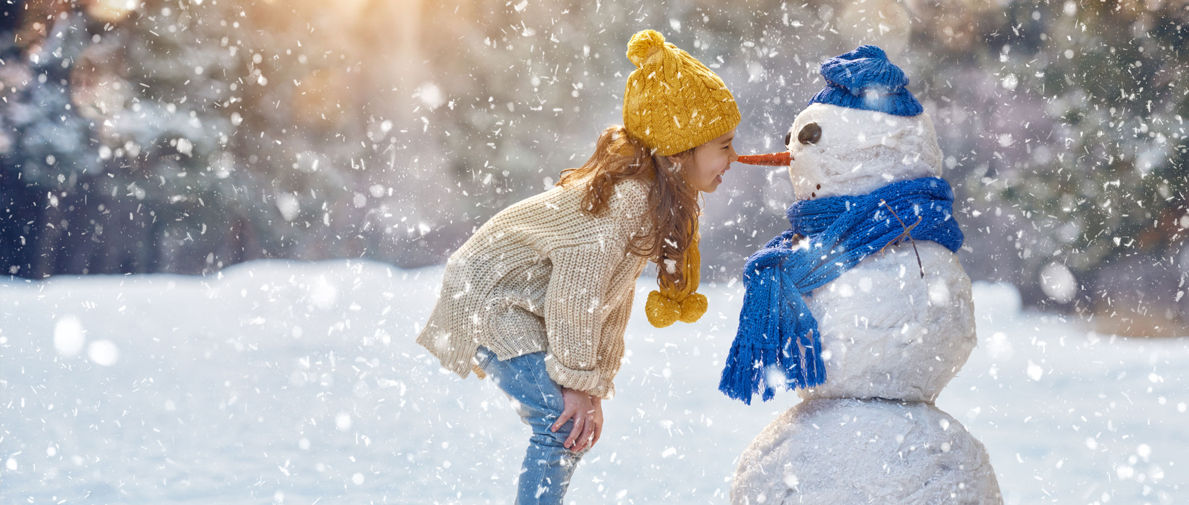 Une fillette portant une tuque et une écharpe jaunes se tient « nez à carotte » avec un bonhomme de neige portant une tuque et une écharpe bleues, tandis qu'il neige autour d'eux.