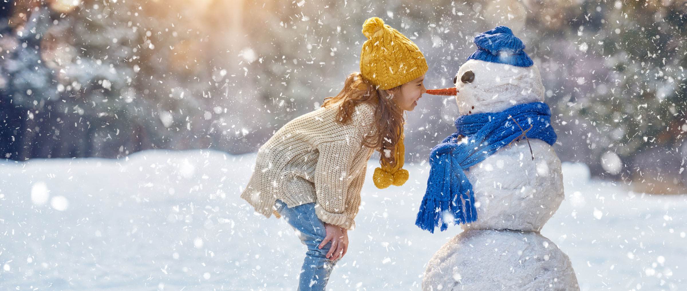 A girl wearing a yellow tuque and scarf stands “nose to carrot” with a snowman that’s wearing a blue tuque and scarf while it snows around them.