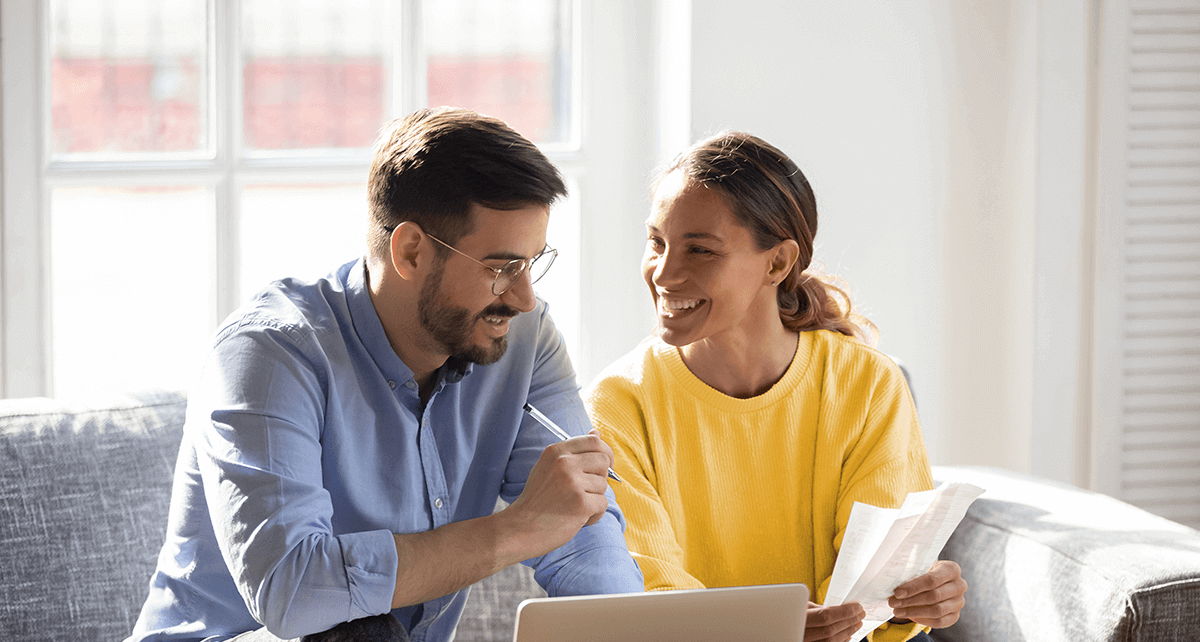 A couple sit together on a couch with a laptop, papers and a calculator on the table in front of them.