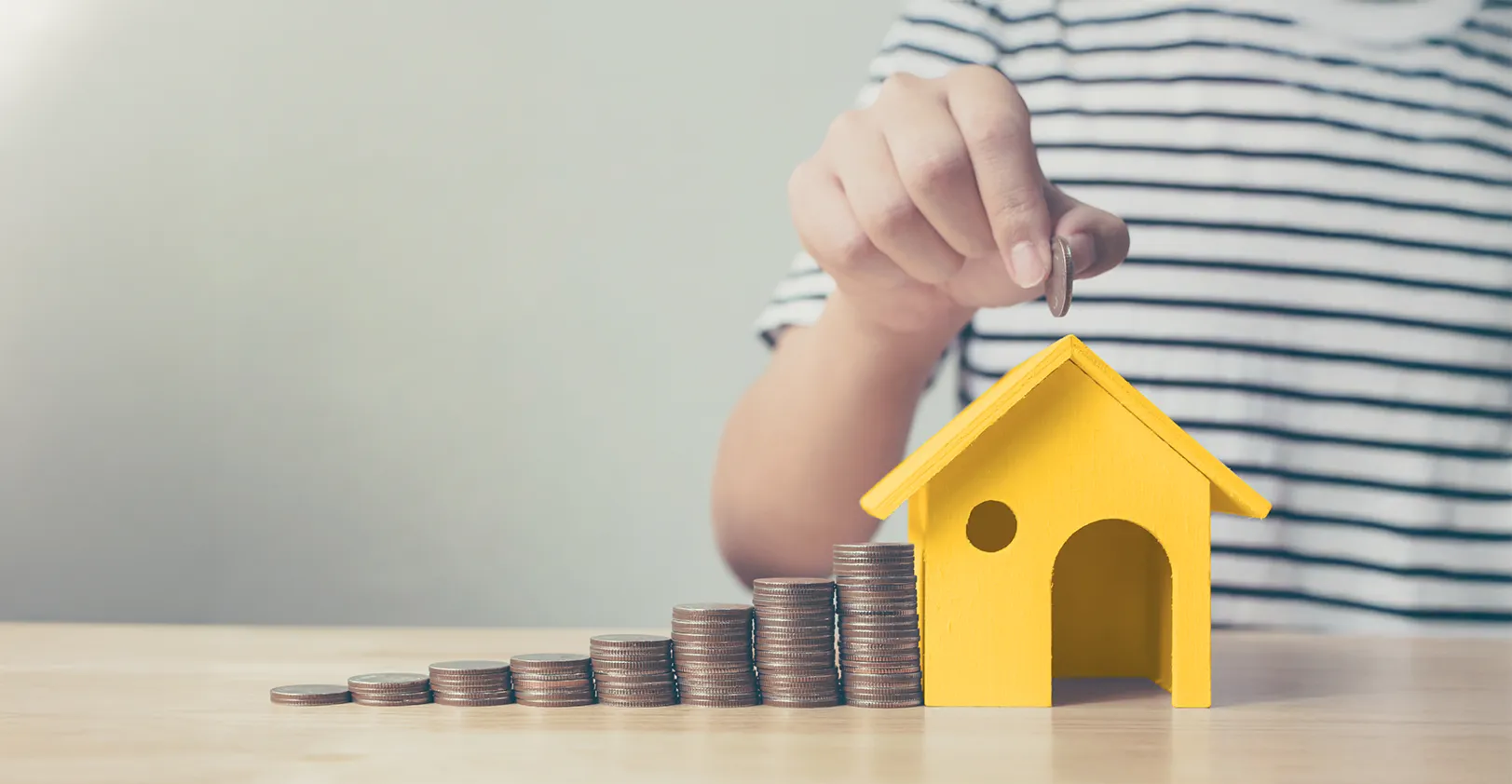 A hand holds a coin above small wooden model of a yellow house. To the left of the house are 8 stacks of coins that steadily get taller.