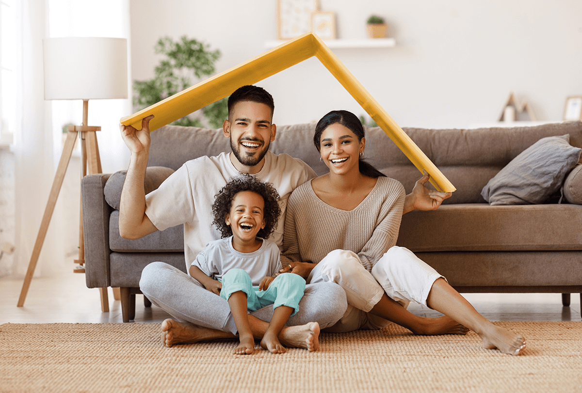 Une jeune famille de trois personnes est assise joyeusement sur le sol de son salon. L'enfant sur les genoux, le père tient les deux côtés d'une boîte au-dessus de la tête de chacun, comme un toit.