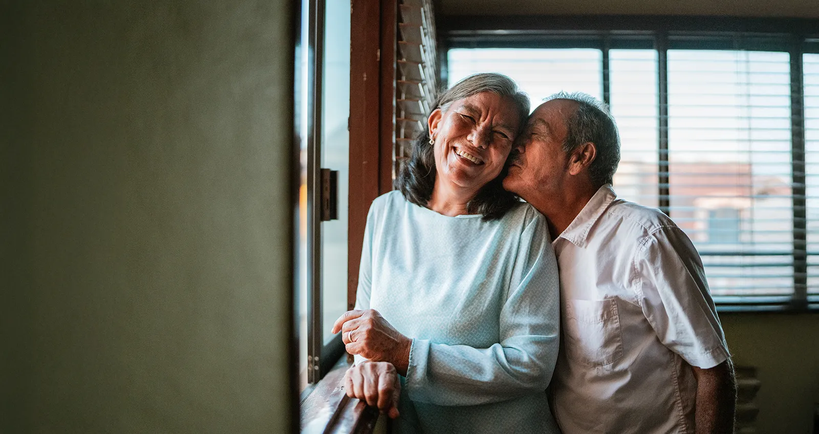 An elderly couple share a moment by a window, the man affectionately kissing the woman on the cheek as she smiles.