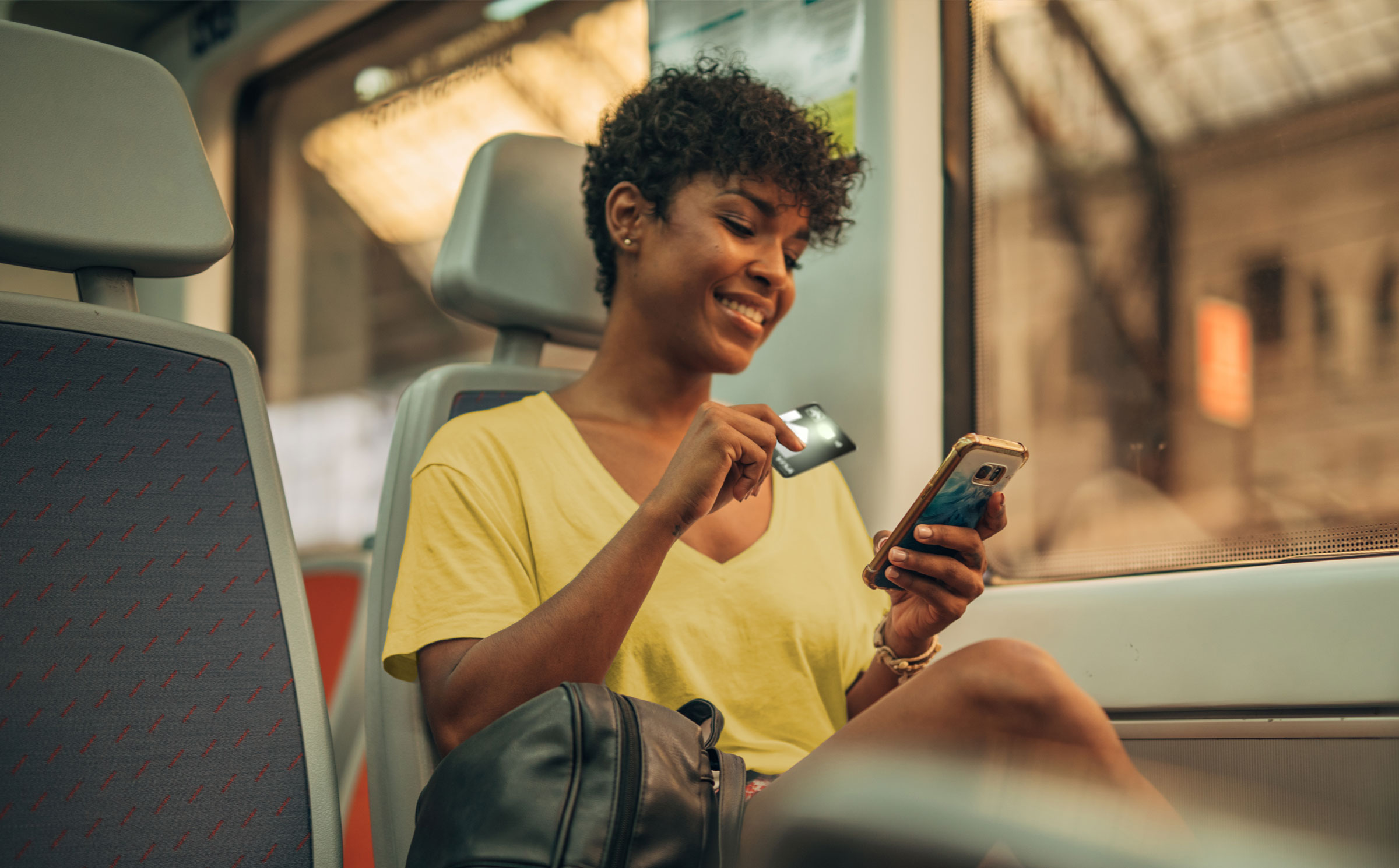 Une femme est assise dans un train. Elle tient sa carte de crédit de la Banque Laurentienne dans une main et son téléphone intelligent dans l’autre. Elle sourit en regardant son téléphone intelligent.