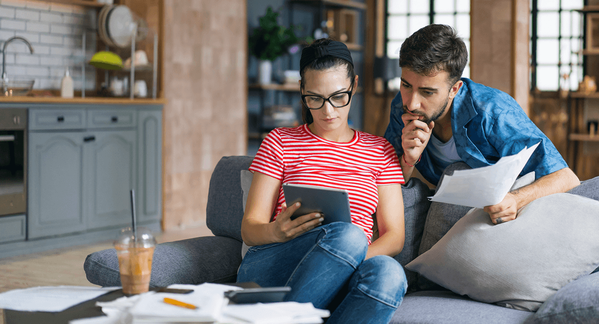 Un couple est assis dans son salon. La femme est assise sur le canapé et tient une tablette. Son partenaire se penche par-dessus son épaule pour regarder la tablette, des papiers à la main.