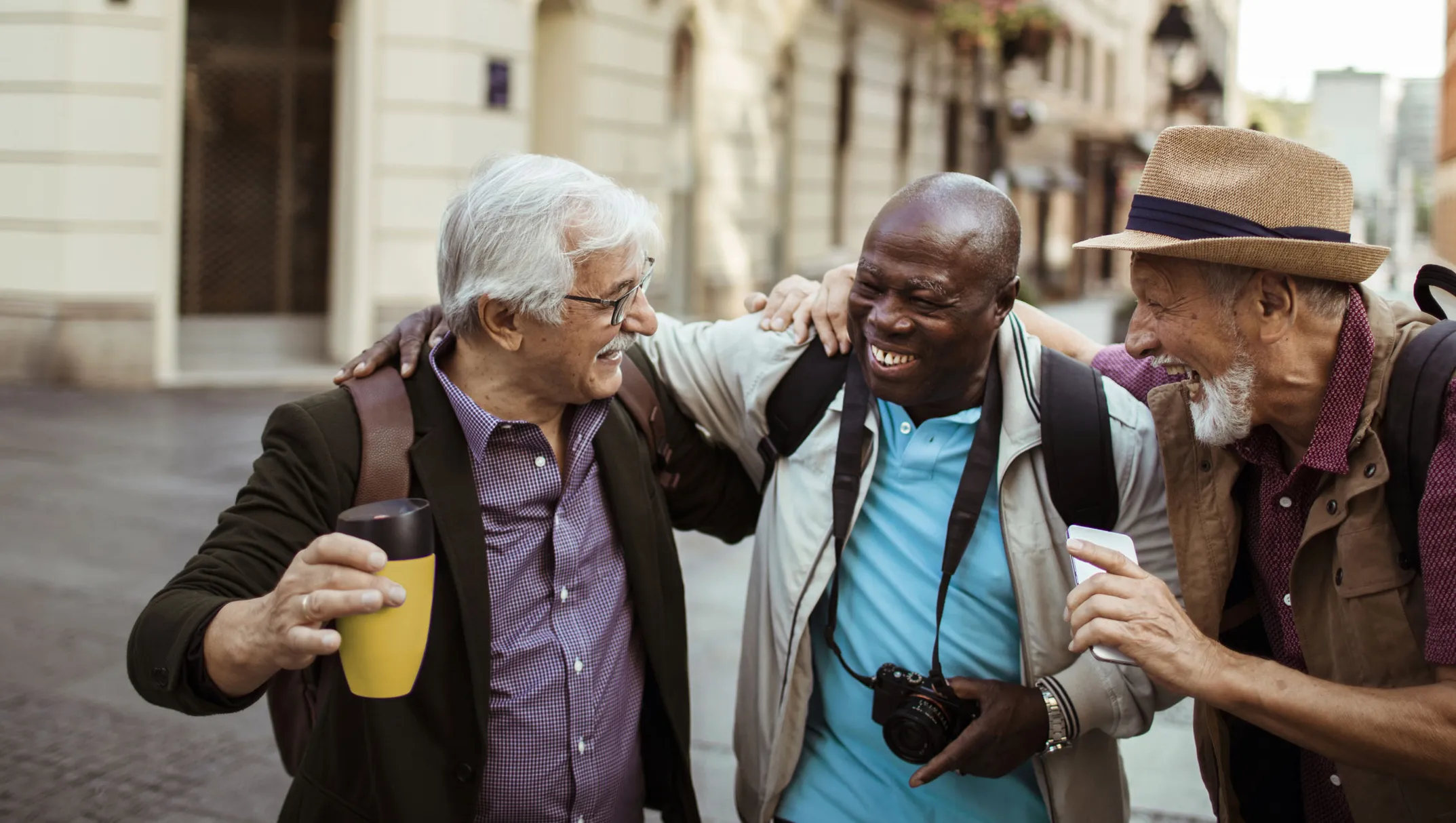 Trois hommes d’un certain âge rient ensemble, bras dessus, bras dessous, dans une rue pittoresque. L'un tient une tasse de voyage, l'autre un appareil photo et le troisième un téléphone mobile.