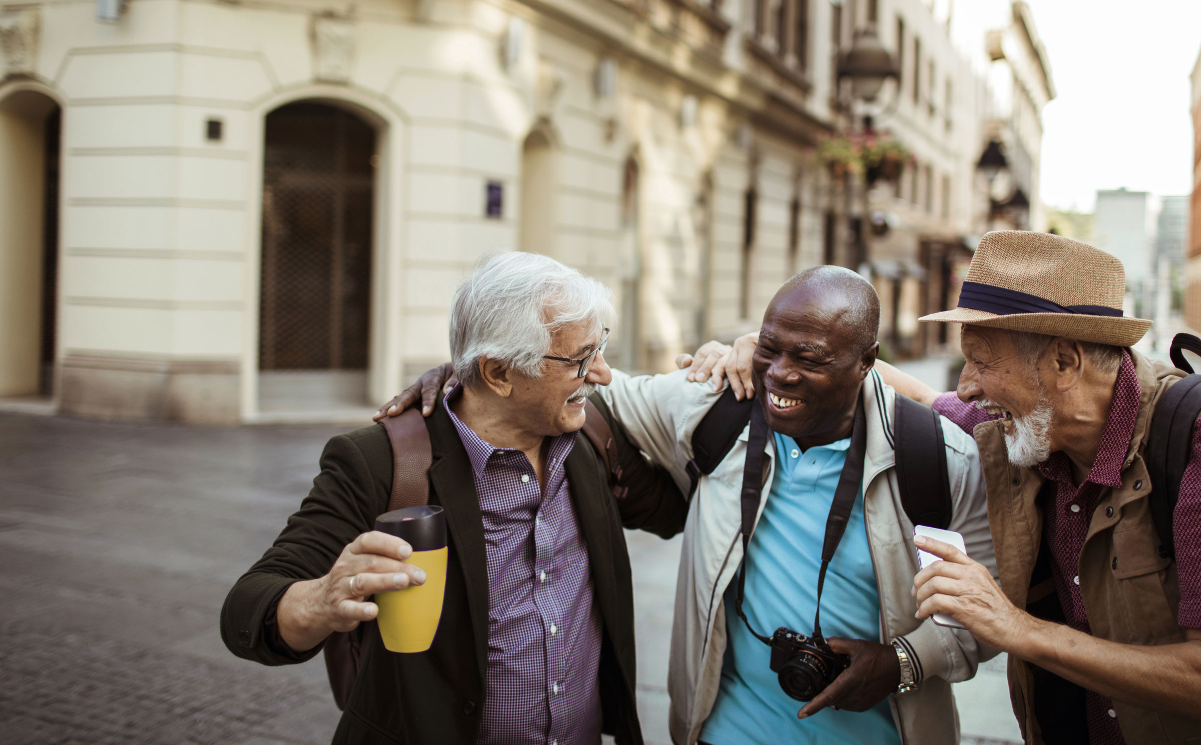 Three older men share a laugh with arms around each other’s shoulders while on a picturesque street. One holds a travel mug, one a camera, the other a cell phone.