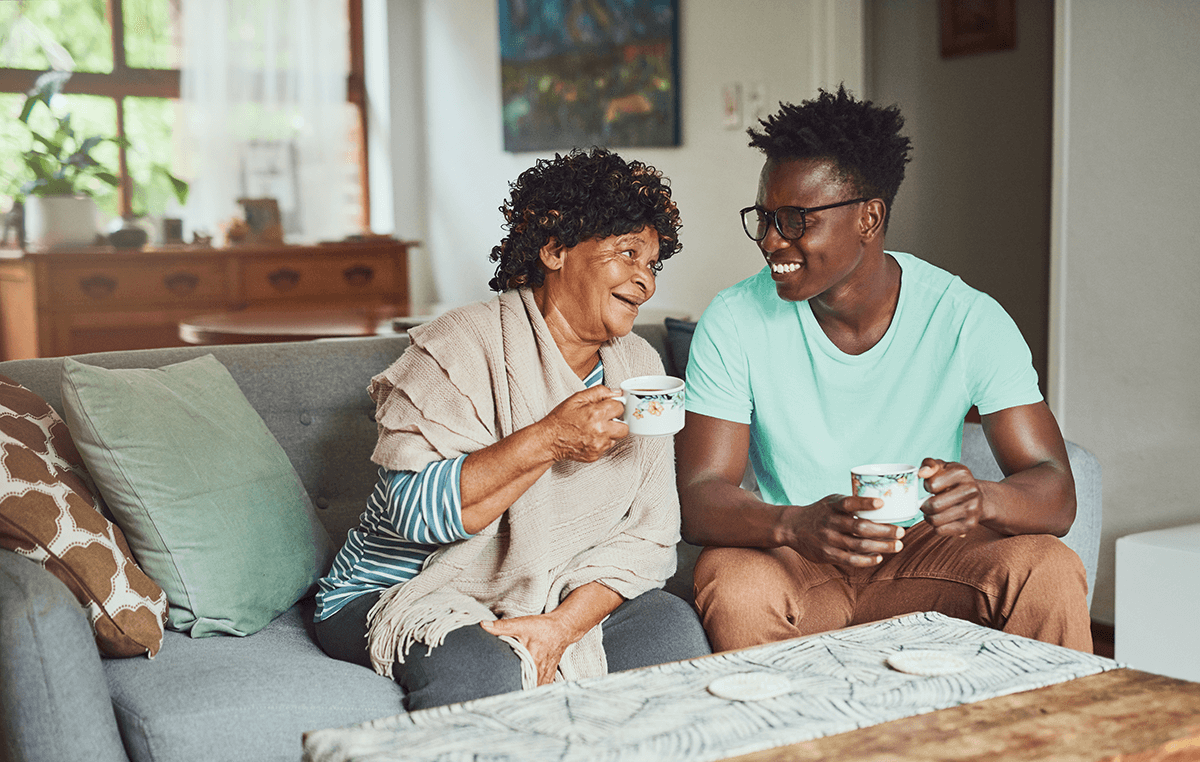 A grandmother and grandson sit together on a sofa, smiling at one another while they each hold a cup of tea.