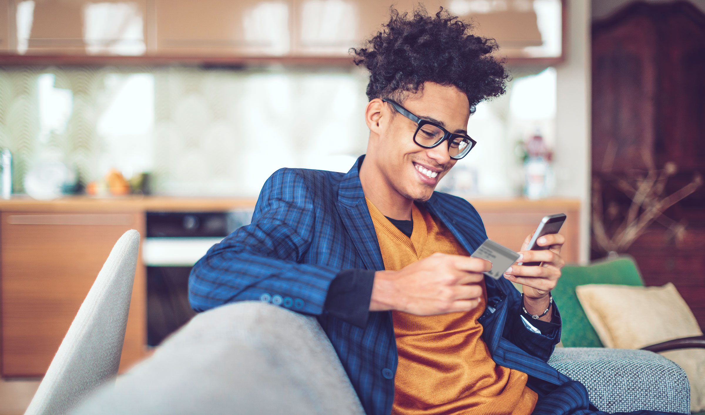 Un jeune homme est assis sur un canapé, tenant une carte de crédit dans une main et un téléphone mobile dans l'autre. Il sourit en regardant son téléphone.